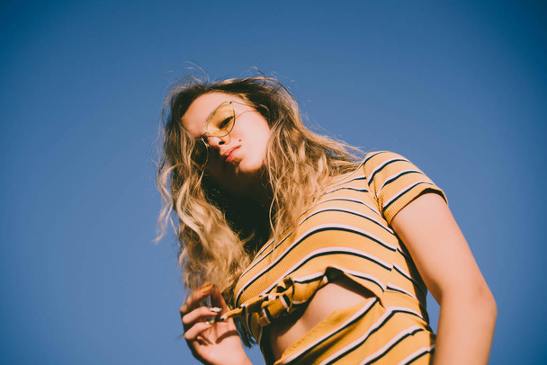 adolescente feliz menina atraente olha para a câmera na praia ensolarada sobre o céu azul usando óculos de sol amarelos
. - Foto, Imagem
