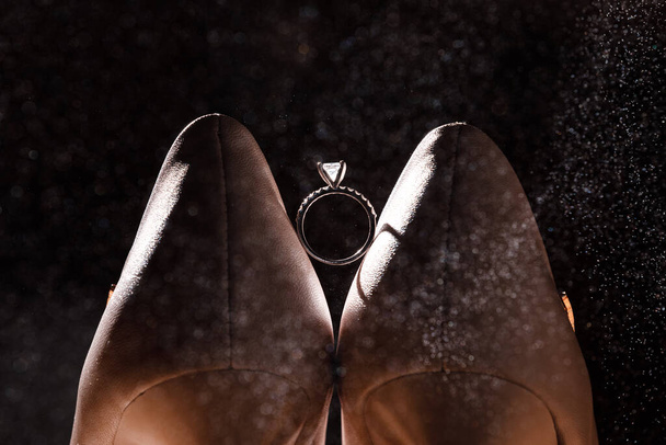 Золотое кольцо с бриллиантом между парой белых туфель на темном фоне
 - Фото, изображение