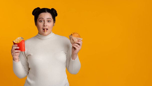 Υπερβολική νεαρή γυναίκα με τηγανητές πατάτες σερβιρίσματος και burger στα χέρια - Φωτογραφία, εικόνα