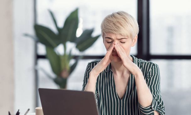 Γυναίκα που αγγίζει τη μύτη έχοντας πόνο κόλπων κάθεται στο χώρο εργασίας εσωτερικούς χώρους - Φωτογραφία, εικόνα