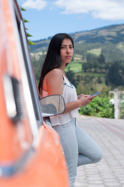 portrait of girl with cellphone in hands dressed in white blouse and jeans sitting in a classic tomato car - Photo, Image
