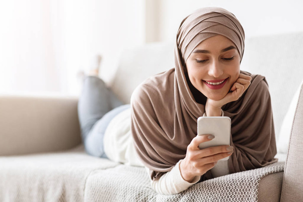 Arap kız evde rahat bir kanepede uzanırken akıllı telefon kullanıyor. - Fotoğraf, Görsel