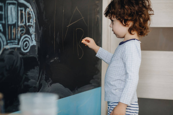 Curly little boy draws chalk on a board in his room - Фото, изображение