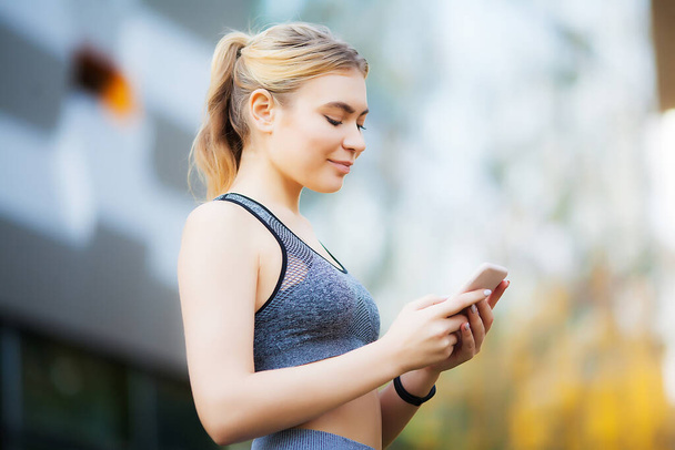 Φωτογραφία της γυναίκα χαρούμενη Fitness 30s Sportswear αγγίζοντας Bluetooth Earpod και κρατώντας το κινητό τηλέφωνο, ενώ αναπαύεται στο Green Park - Φωτογραφία, εικόνα