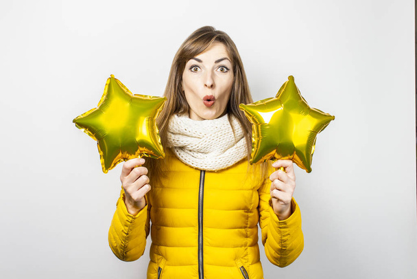 Woman with a surprised face in a yellow jacket and a scarf holding star air balloons on a white background. Concept of a holiday, celebration, party, new year. Banner - Photo, Image