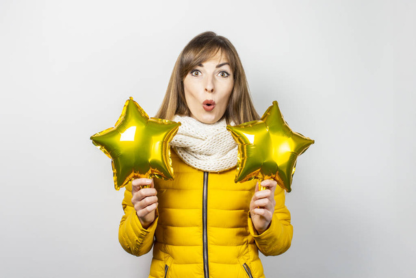 Mujer con una cara sorprendida con una chaqueta amarilla y una bufanda que sostiene globos de aire estrellados sobre un fondo blanco. El concepto de la fiesta, la fiesta, la fiesta, un nuevo año. Banner - Foto, Imagen