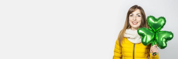 Woman with a smile in a yellow jacket and a scarf holds a clover balloon on a white background. Concept of a holiday, celebration, party, St. Patrick's Day, Ireland. Banner - 写真・画像