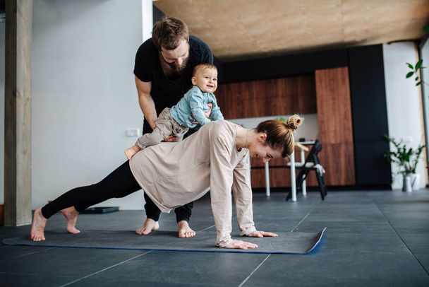 Family morning exercise. Mother doing plank, father holding baby on her back - Photo, Image
