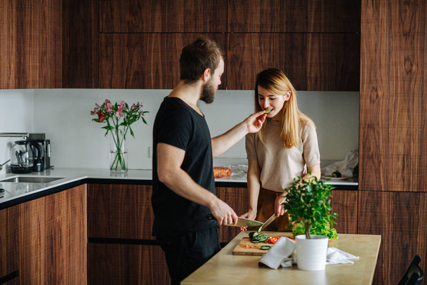 Woman cutting vegetables, while her boyfriend feeding her cucumber slices - Photo, image