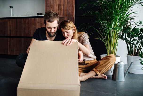 Young man and woman sitting on a floor, peeking inside a box. - Zdjęcie, obraz
