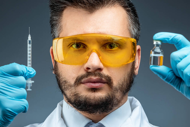 Чоловік-вчений, лабораторний асистент у білому пальто та окулярах тримає пляшку з вакциною в руках, медичні тести, лабораторії, експерименти. Хімічний аналіз імунізації
. - Фото, зображення