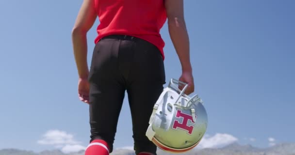 Low angle rear view of a mixed-raced American football player holding his helmet and walking away from camera, against a blue sky at a sports field, in slow motion. Track and Field Sports Training in Stadium. - Filmagem, Vídeo