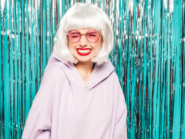 Young sexy smiling hipster girl in white wig and red lips.Beautiful trendy woman in summer clothes.Carefree model posing on blue silver shiny tinsel background in studio.Positive female going crazy - Foto, Bild