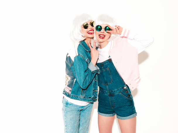 Two young sexy smiling hipster girls in white wigs and red lips.Beautiful trendy women in summer jeans clothes.Carefree models isolated on white in studio.Positive female shows tongue and peace sign - Fotografie, Obrázek