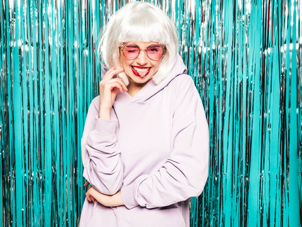 Young sexy smiling hipster girl in white wig and red lips.Beautiful trendy woman in summer clothes.Carefree model posing on blue silver shiny tinsel background in studio.Positive female shows tongue and peace sign - Foto, imagen