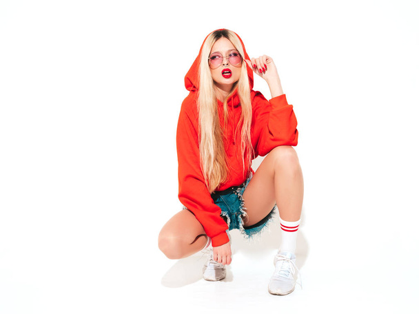 Portrait of young beautiful hipster bad girl in trendy red summer hoodie and earring in her nose.Sexy carefree smiling blond woman sitting in studio.Positive model having fun.Isolated on white - Foto, imagen