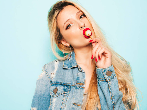 Portrait of young beautiful hipster bad girl in trendy jeans summer clothes and earring in her nose.Sexy carefree smiling blond woman posing in studio.Positive model licking round sugar candy - Foto, Bild
