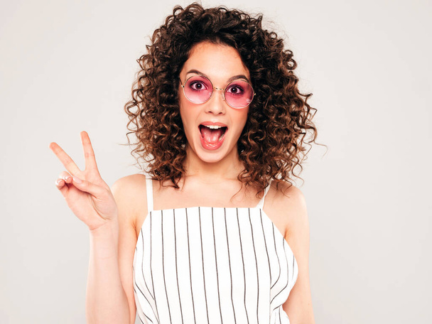 Portrait of beautiful smiling model with afro curls hairstyle dressed in summer hipster clothes.Sexy carefree girl posing in studio on gray background.Trendy funny and positive woman shows peace sign - 写真・画像