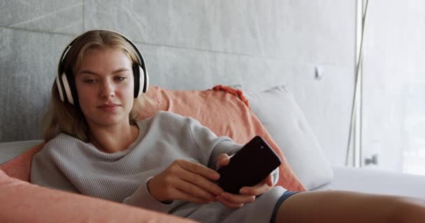 Vista frontal de una mujer caucásica disfrutando de un tiempo de calidad en un hotel, acostada en la cama en el dormitorio, escuchando música y usando un teléfono inteligente, cámara lenta
 - Metraje, vídeo