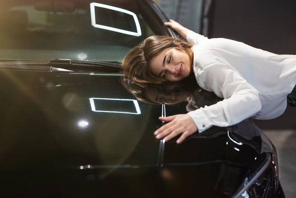young beautiful woman feels happy and joyful hugging her brand new vehicle in electric car dealership center, close up - Фото, зображення