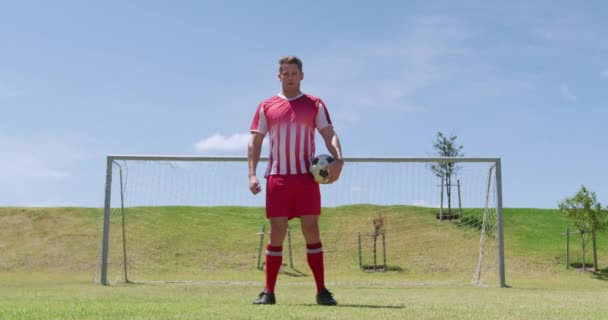 Front view of Caucasian male football player wearing a team strip, training at a sports field standing in front of a goal holding a football and looking to camera, in slow motion. Track and Field Sports Training in Stadium. - Záběry, video