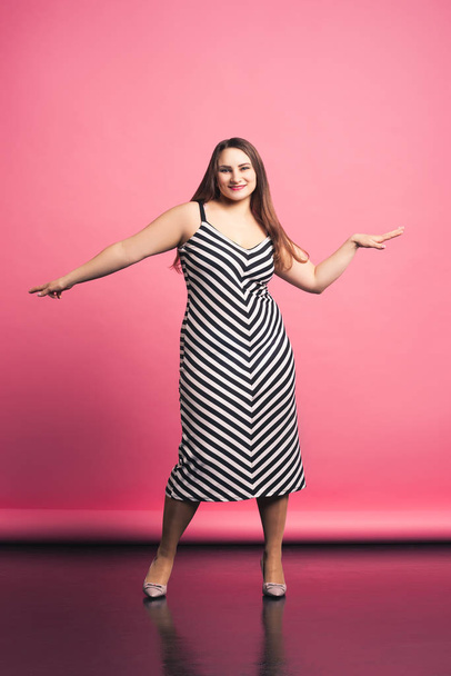 Happy pize size model in striped dress, fat woman dancing on pink background - Foto, afbeelding