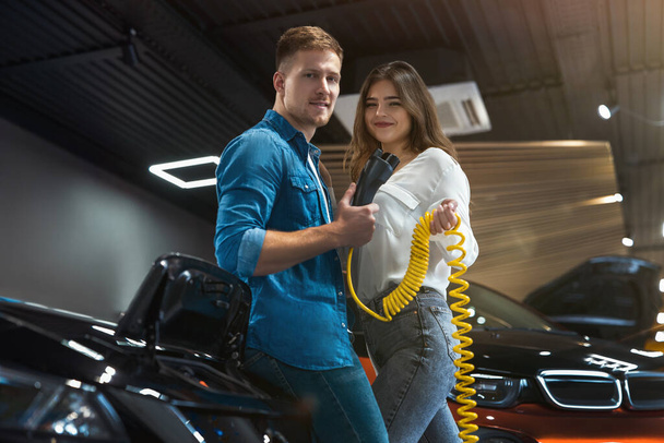 handsome man and beautiful brunette woman hold charging cable for electric car standing near open bonnet in the electric vehicle dealership center , eco friendly concept - Fotografie, Obrázek