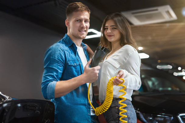 handsome man and beautiful brunette woman hold charging cable for electric car standing in the electric vehicle dealership center , eco friendly concept - Fotografie, Obrázek