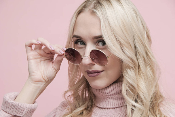 Happy coquettish woman face with blond hair in glasses and wool sweater on pastel pink background. Close up - Foto, Bild