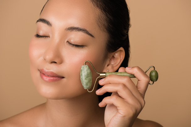 smiling beautiful naked asian girl with closed eyes massaging face with jade roller isolated on beige - Foto, imagen