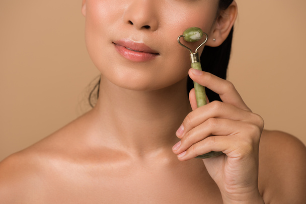 recortada vista de hermosa chica asiática desnuda masajeando cara con rodillo de jade aislado en beige
 - Foto, Imagen
