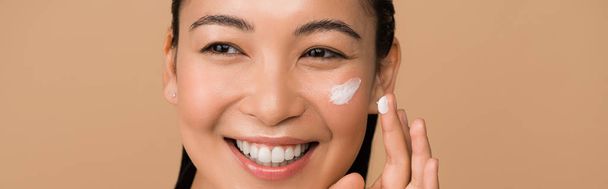 felice bella ragazza asiatica applicando crema per il viso isolato su beige, colpo panoramico
 - Foto, immagini