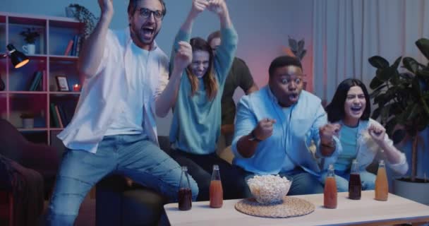 Happy guys and girls celebrate winning of team while sitting on sofa at home. Group of excited friends rejoicing victory while watching sport games on TV. Concept of sport and emotions. - Metraje, vídeo