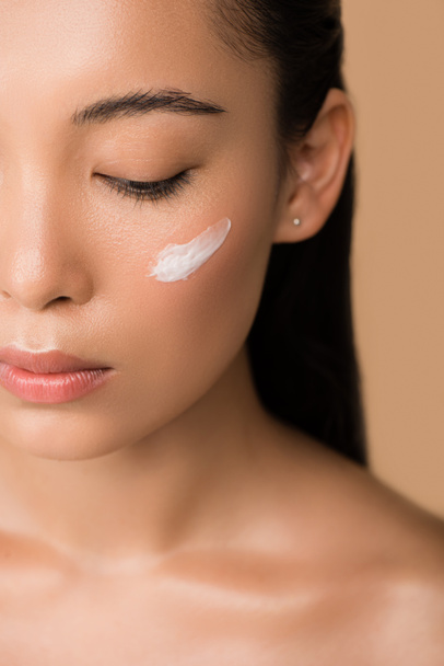 cropped view of beautiful naked asian girl with facial cream on cheek isolated on beige - Foto, Imagen