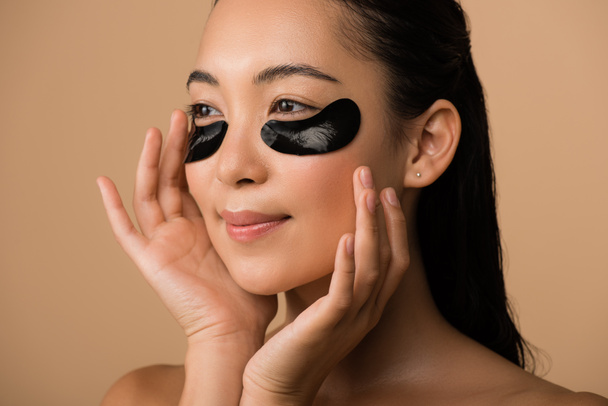 smiling beautiful naked asian girl with black hydrogel eye patches under eyes isolated on beige - Photo, Image