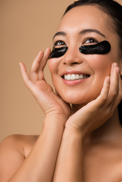 χαμογελαστή όμορφη γυμνή Ασιάτισσα κοπέλα με μαύρα μπαλώματα υδρογέλης κάτω από τα μάτια απομονωμένη σε μπεζ - Φωτογραφία, εικόνα