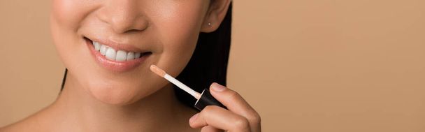 cropped view of smiling beautiful asian girl appying lip gloss on lips isolated on beige, panoramic shot - Foto, Imagem