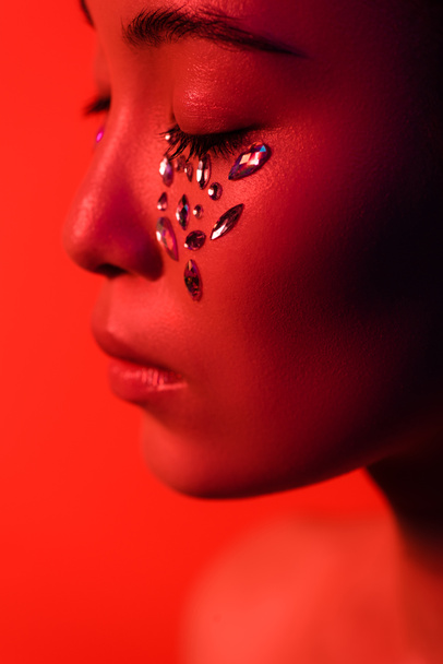 profil pięknej azjatyckiej dziewczyny z kryształkami na twarzy i zamkniętych oczach odizolowanych na czerwono - Zdjęcie, obraz