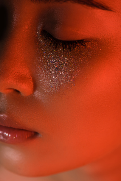 close up view of beautiful asian girl with silver sparkles on face and closed eyes in red lighting - Foto, Imagem