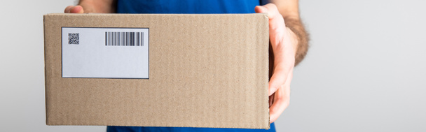 Cropped view of delivery man holding cardboard box with qr code and barcode isolated on grey, panoramic shot - Foto, Imagem