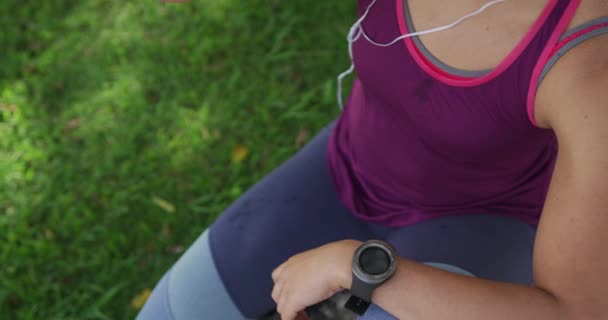 High angle mid section view of a young Caucasian woman wearing sports clothes leaning sitting, drinking water and looking at watch during a workout in a park, slow motion - Video, Çekim