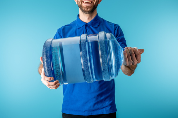 Vágott kilátás mosolygós futár gazdaság palackozott víz elszigetelt kék - Fotó, kép