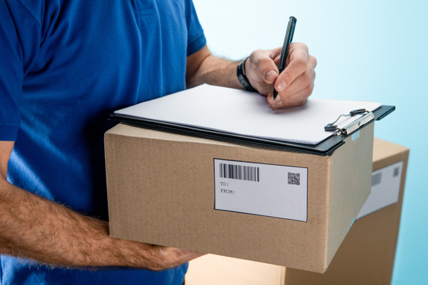 Cropped view of courier writing on clipboard on cardboard box with packages isolated on blue - Foto, afbeelding