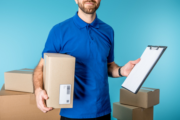 Oříznutý pohled na doručovatele držícího lepenkovou krabici a schránku s kopírovacím prostorem v blízkosti krabic izolovaných na modré - Fotografie, Obrázek