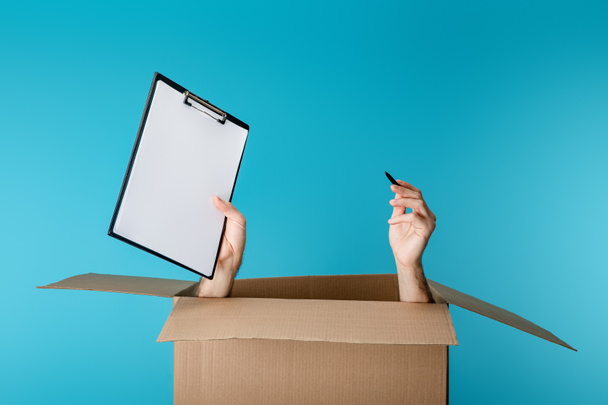 Руки курьера, держащего планшет и ручку из картонной коробки, изолированной на голубом
 - Фото, изображение