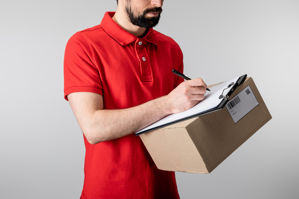 Oříznutý pohled na doručovatele, jak píše na schránku a drží kartonovou krabici izolovanou na šedé - Fotografie, Obrázek