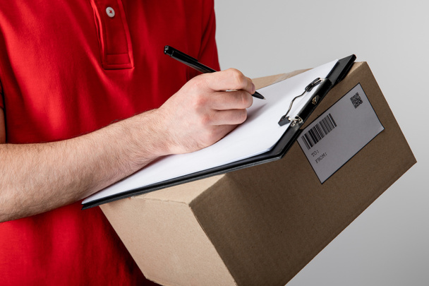 Cropped view of courier writing on clipboard with empty blank and holding cardboard box isolated on grey - Photo, Image