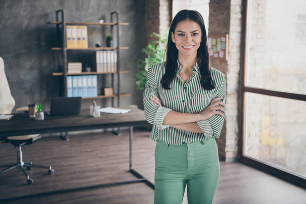 Photo of attractive cheerful latin business lady arms crossed reliable person meet partners friendly smiling assistant wear striped shirt green pants standing modern office indoors - Foto, imagen