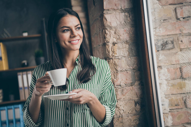 Photo of attractive latin business lady hold white porcelain cup morning espresso coffee look dreamy window lean brick wall wear striped shirt modern office indoors - Foto, Imagen