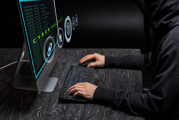 oříznutý pohled na psaní hackerů na klávesnici počítače poblíž monitoru počítače s kybernetickým zabezpečovacím písmem na černé  - Fotografie, Obrázek